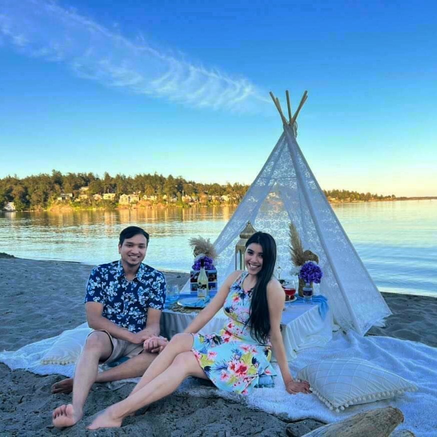 A smiling couple seated on a rug beside a lace teepee beach picnic at sunset