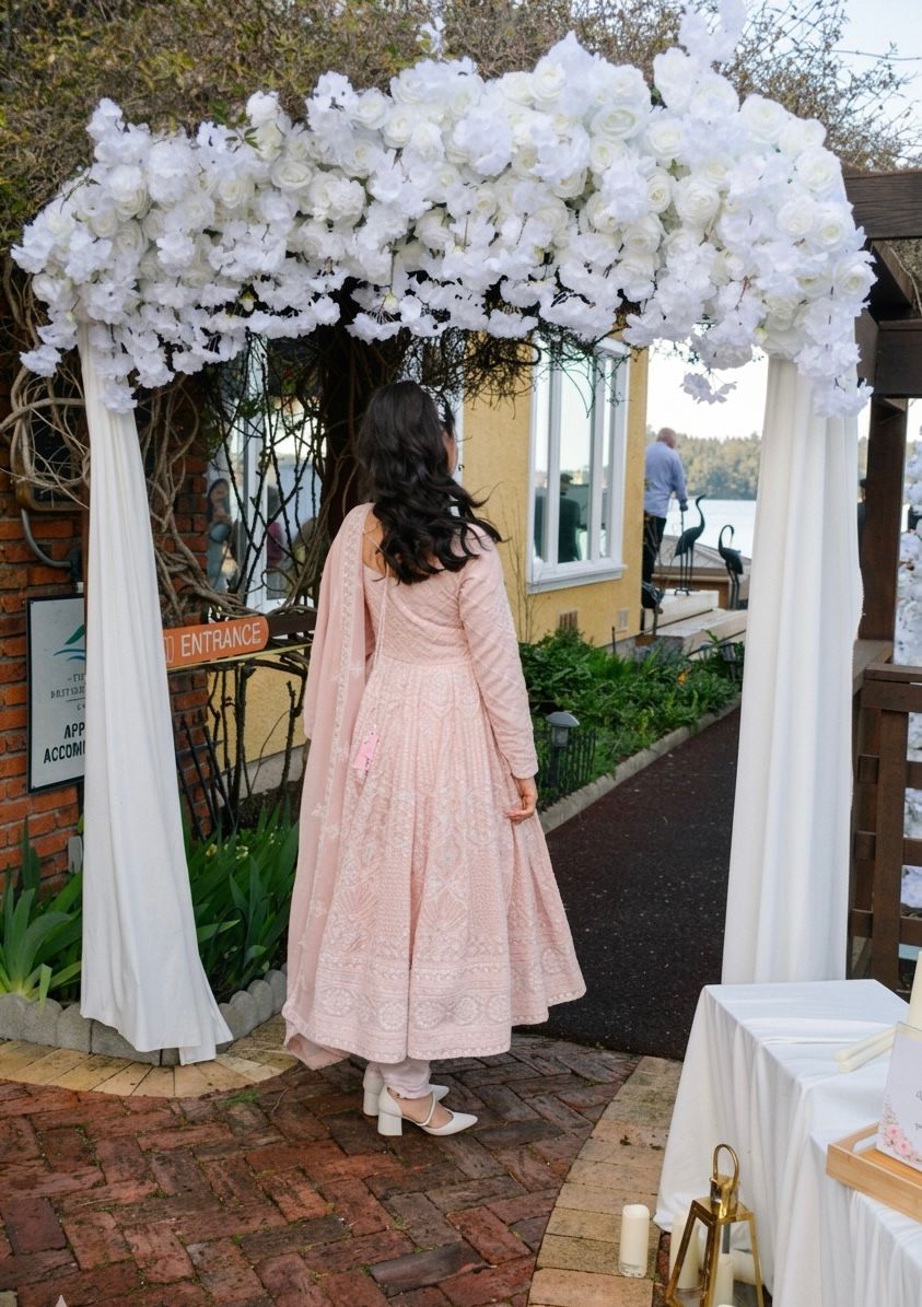 A bride in a pink outfit walking through a full white rose arch by the water