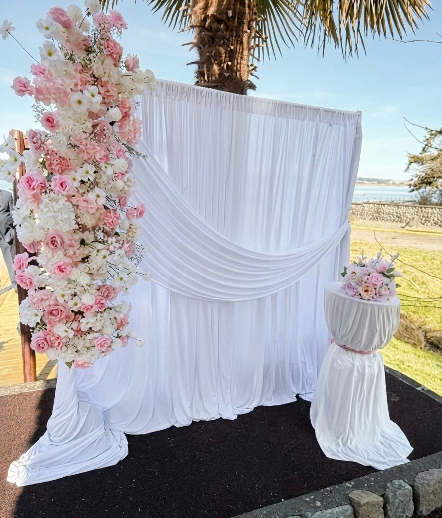 Pink and white floral arch beside a white draped backdrop at a waterfront ceremony
